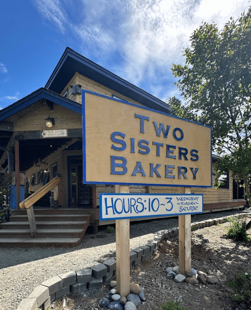 Sign for "Two Sisters Bakery" with hours listed as Wednesday to Saturday, 10 AM to 3 PM, in front of a charming building.