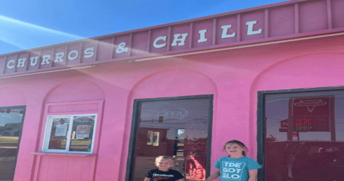 Two children stand in front of a pink building with a sign that reads "Churros & Chill." Bright sunlight shines above.