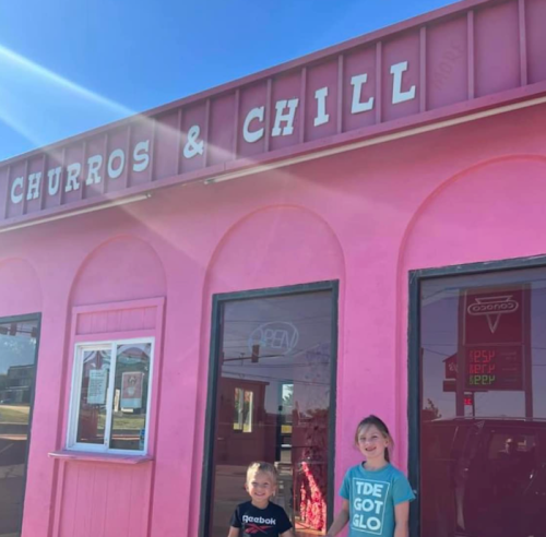 Two children stand in front of a pink building with a sign that reads "Churros & Chill." Bright sunny day.