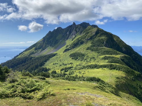 A lush green mountain rises under a blue sky with scattered clouds, overlooking a distant ocean.