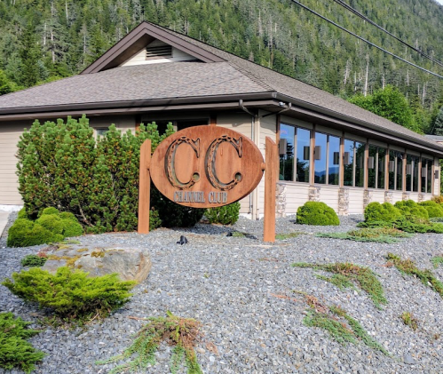 Sign for "C.C. Cannery Club" in front of a building surrounded by greenery and gravel, with mountains in the background.