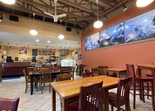 Cozy café interior with wooden tables, chairs, and wall art, featuring a counter and display case in the background.