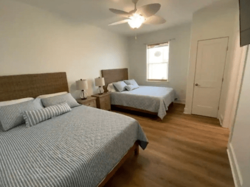 A bright bedroom featuring two beds, bedside tables, lamps, and a ceiling fan, with light wood flooring.