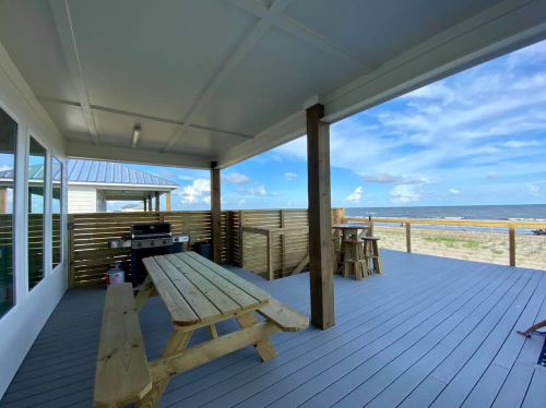 A spacious outdoor deck with a picnic table, grill, and ocean view under a blue sky with scattered clouds.