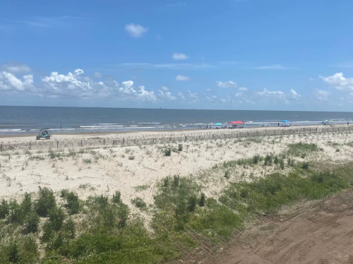 A sandy beach with sparse vegetation, umbrellas, and a car parked near the shoreline under a blue sky with clouds.