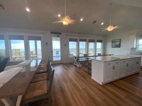 Bright, open kitchen and dining area with large windows overlooking a beach view, featuring modern decor and wooden floors.