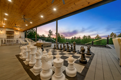 A large outdoor chess set on a deck with a sunset view and modern seating in the background.