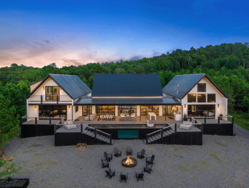 A modern two-story house surrounded by trees, featuring a fire pit and outdoor seating area at dusk.