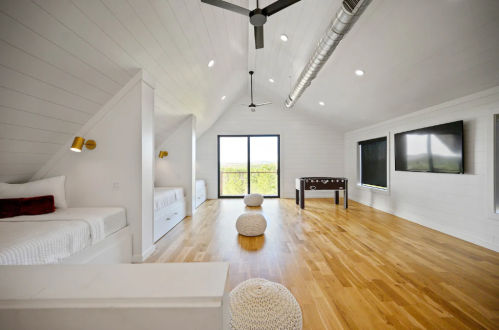 Bright, modern room with two beds, a foosball table, and large windows overlooking greenery. Wood flooring and white walls.