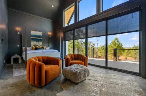 Modern bedroom with large windows, featuring a bed, two orange chairs, and a cozy rug, overlooking a natural landscape.