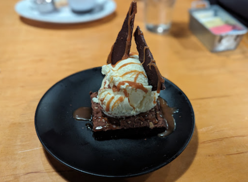 A chocolate brownie topped with vanilla ice cream, caramel drizzle, and chocolate shards on a black plate.