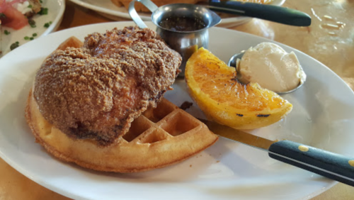 A plate featuring a crispy fried chicken breast on a waffle, with a slice of grilled orange and a small cup of syrup.