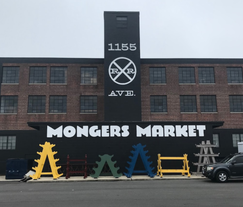 A brick building with "Mongers Market" painted on it, featuring colorful display racks in front.
