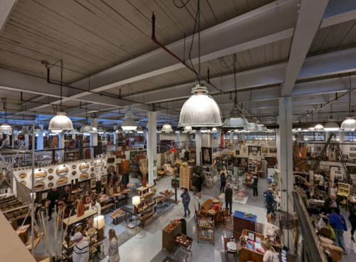 A spacious vintage market filled with various furniture and decor items, with shoppers browsing among the displays.