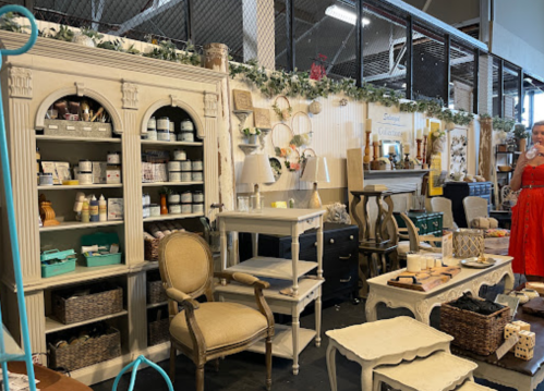 A vintage-style shop display featuring furniture, decor, and various home goods, with a person in a red dress browsing.