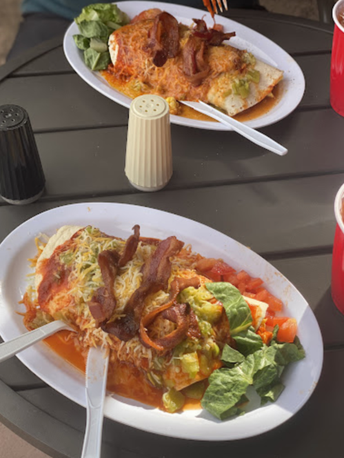 Two plates of enchiladas topped with cheese and bacon, served with lettuce and tomatoes, on a table with condiments.