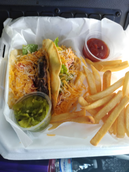 A takeout container with two tacos topped with cheese and lettuce, served with fries and two dipping sauces.