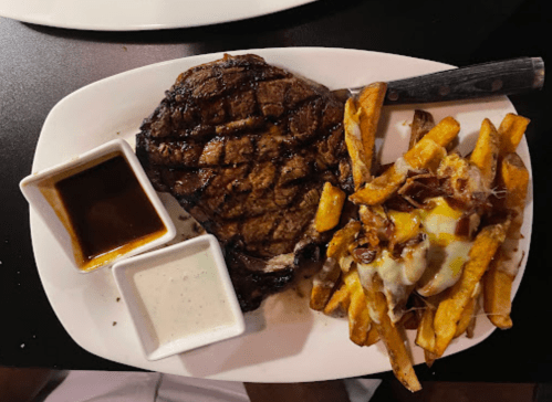 A grilled steak with a side of loaded fries topped with cheese and bacon, served with two dipping sauces.
