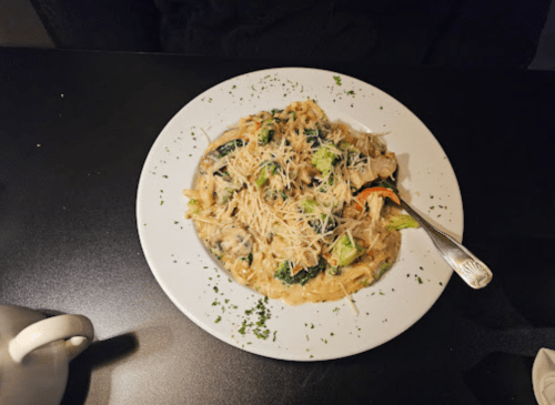 A plate of creamy pasta with vegetables and grated cheese, garnished with parsley, on a dark table.