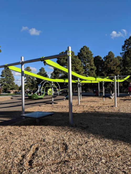 A playground with green canopies, metal structures, and a sandy ground, surrounded by trees and a blue sky.