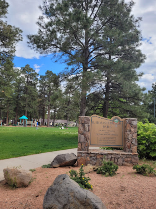 Sign for Bushmaster Park surrounded by trees, with a grassy area and playground visible in the background.