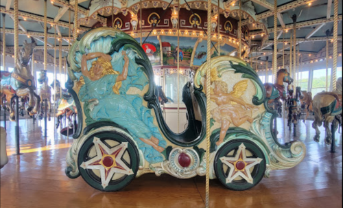 A beautifully crafted carousel chariot featuring intricate sculptures of a reclining figure and an angel, surrounded by horses.