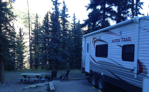 A camper parked in a forested area with picnic table and chairs nearby, surrounded by tall evergreen trees.