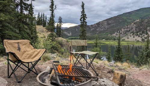 A cozy campsite with two folding chairs by a fire pit, overlooking a serene lake and mountains in the background.