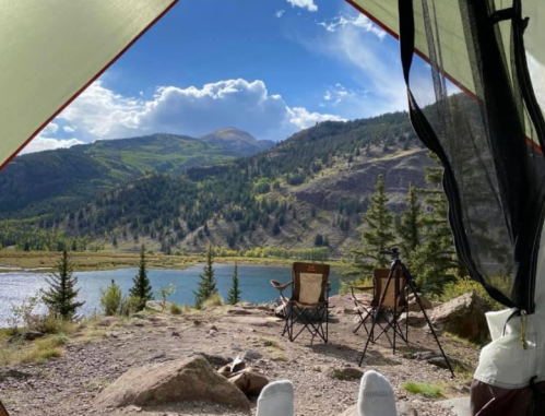 View from inside a tent, showcasing a serene lake and mountains under a blue sky with scattered clouds.