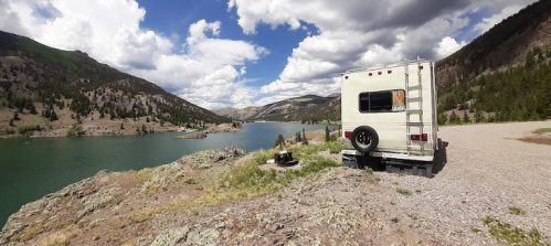A camper van parked by a serene lake, surrounded by mountains and lush greenery under a partly cloudy sky.