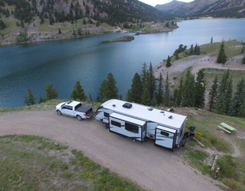 Aerial view of a camper and truck parked by a serene lake surrounded by mountains and trees.