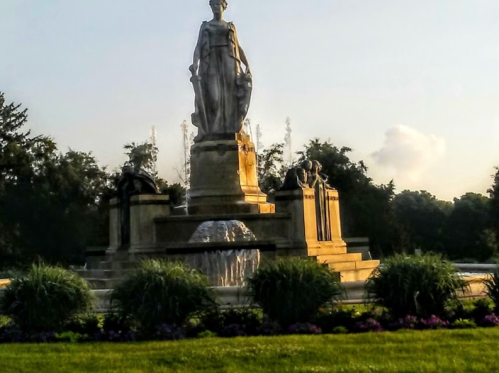 A statue stands atop a fountain, surrounded by lush greenery and colorful flowers in a serene park setting.