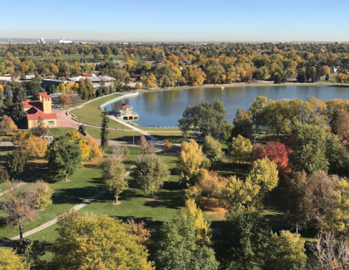 Aerial view of a park with colorful autumn trees surrounding a calm lake and a small building in the background.