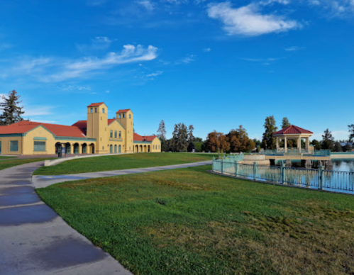 A scenic view of a park featuring a yellow building, gazebo by the water, and a clear blue sky.