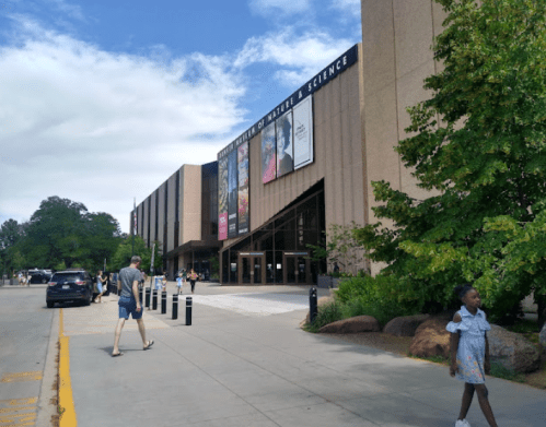 A modern building with large banners, surrounded by trees, people walking, and a clear blue sky.