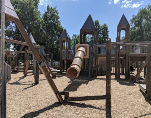 A playground with wooden structures, towers, and a large tube slide, surrounded by wood chips and trees.