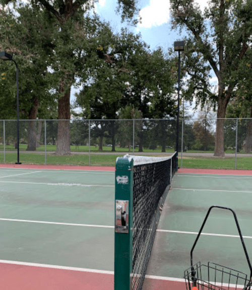A tennis court with a net, surrounded by trees and a cloudy sky, showing empty playing areas.