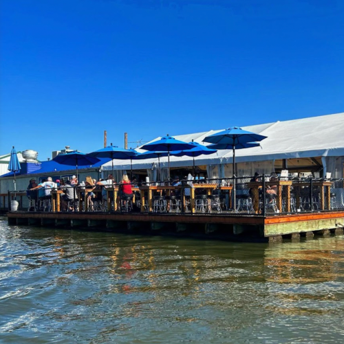 A waterfront restaurant with blue umbrellas, people dining on a deck, and a clear blue sky above.