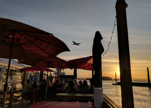 Silhouette of a person by the water at sunset, with an airplane flying overhead and people dining under umbrellas.