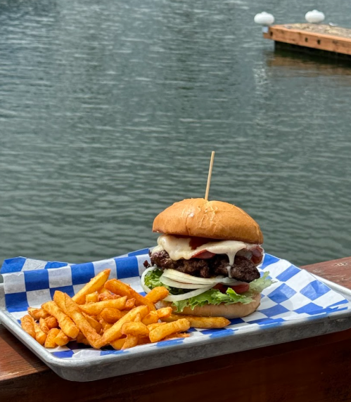 A juicy burger with lettuce, tomato, and onion, served with crispy fries, set against a scenic waterfront background.