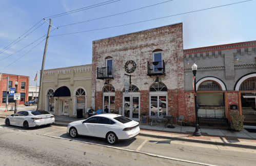 Historic brick building with a clock, featuring storefronts and parked cars on a sunny street.