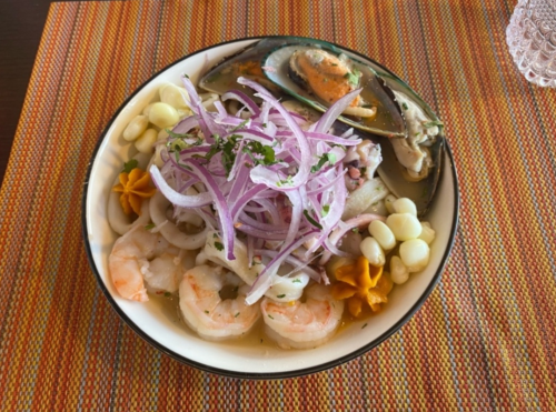 A colorful seafood dish featuring shrimp, mussels, octopus, and garnished with red onions and herbs on a patterned placemat.