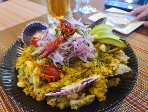 A vibrant seafood dish with rice, shellfish, crawfish, avocado, and onions, served on a black plate.