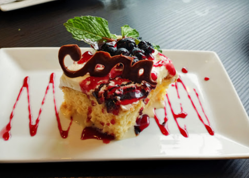 A slice of cake topped with berries, cream, and chocolate decoration, drizzled with raspberry sauce on a white plate.