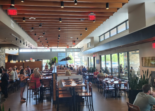 A bustling restaurant interior with wooden beams, tables filled with diners, and large windows letting in natural light.
