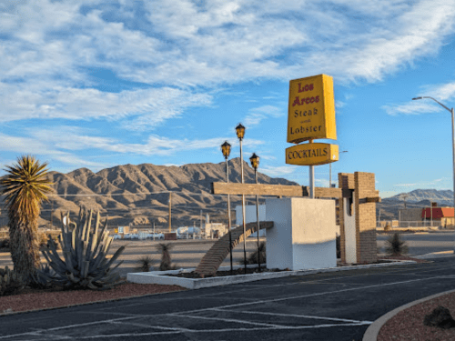 Sign for Los Arcos Steak & Lobster with mountains in the background and palm trees in the foreground. Clear blue sky.