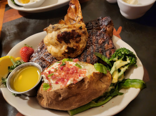 A plate featuring grilled steak, a baked potato with toppings, broccoli, and a strawberry, garnished with lemon and butter.