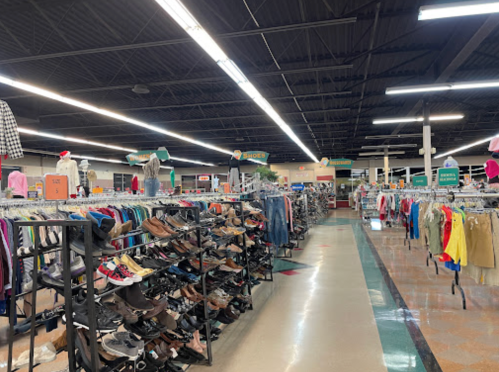 A spacious thrift store aisle filled with clothing racks and shelves of shoes under bright overhead lights.