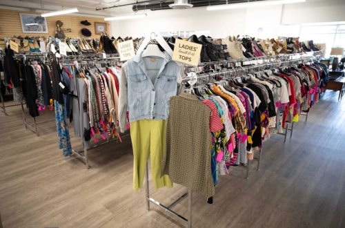 A clothing rack filled with various ladies' tops and pants in a bright, organized thrift store setting.