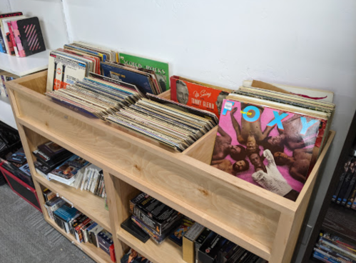 A wooden shelf displaying various vinyl records and albums, with a few visible titles on the front.